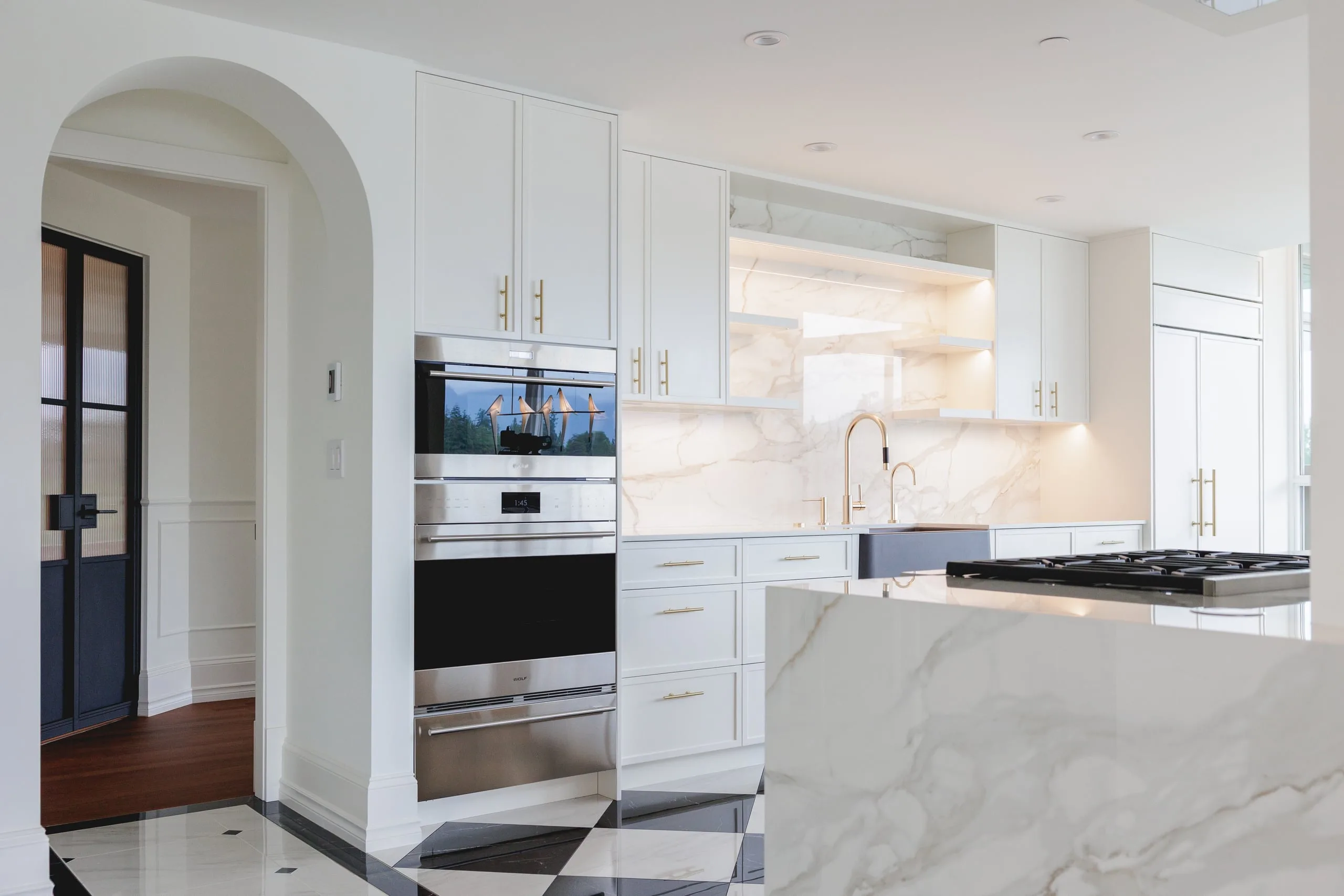 A renovated condo kitchen seen through an arched doorway: wall ovens and a waterfall marble island.