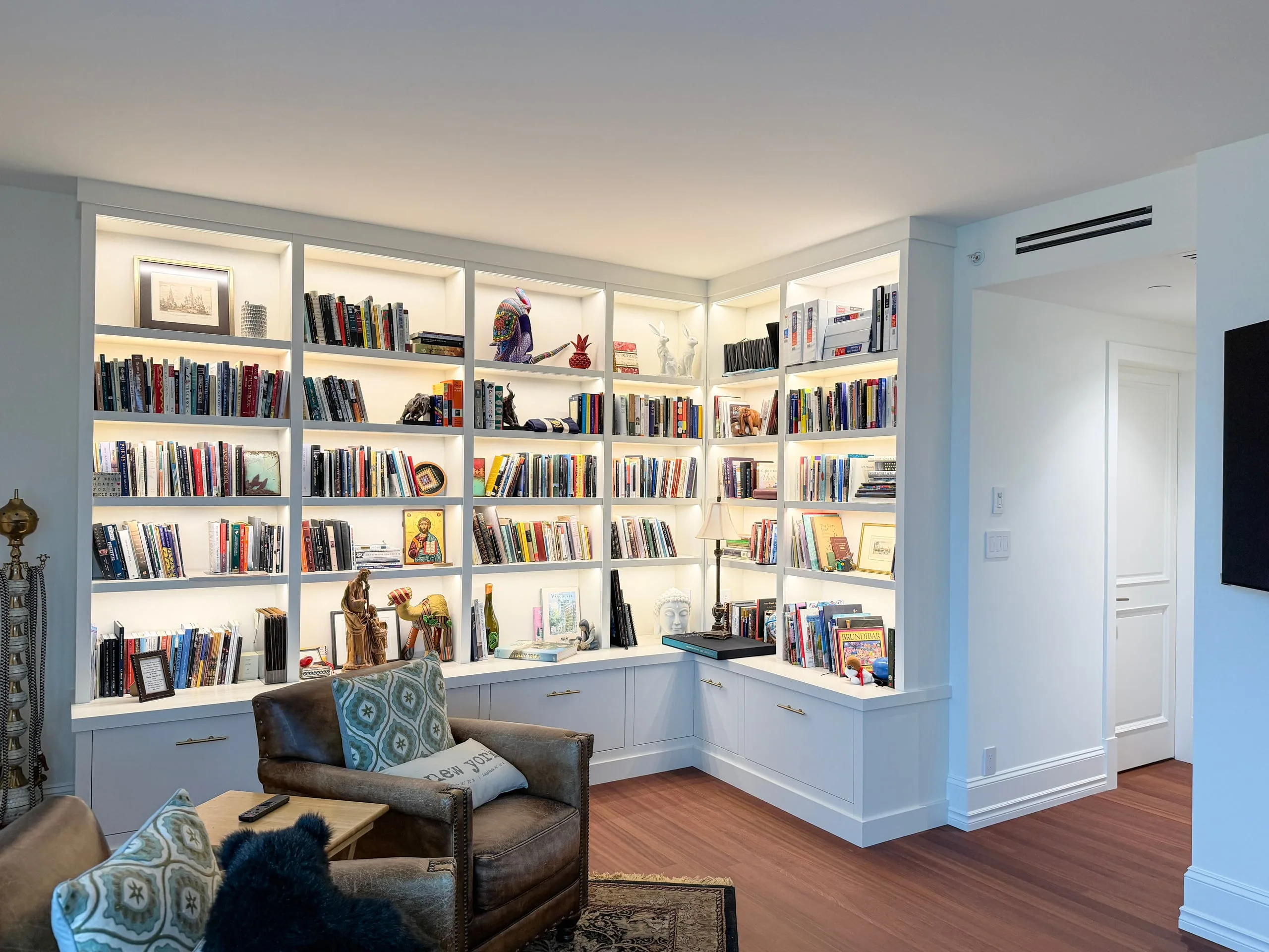 The custom library in The Harbourview Residence, a Coal Harbour condo in Vancouver, with lit white built-in shelves over drawers and a leather armchair.