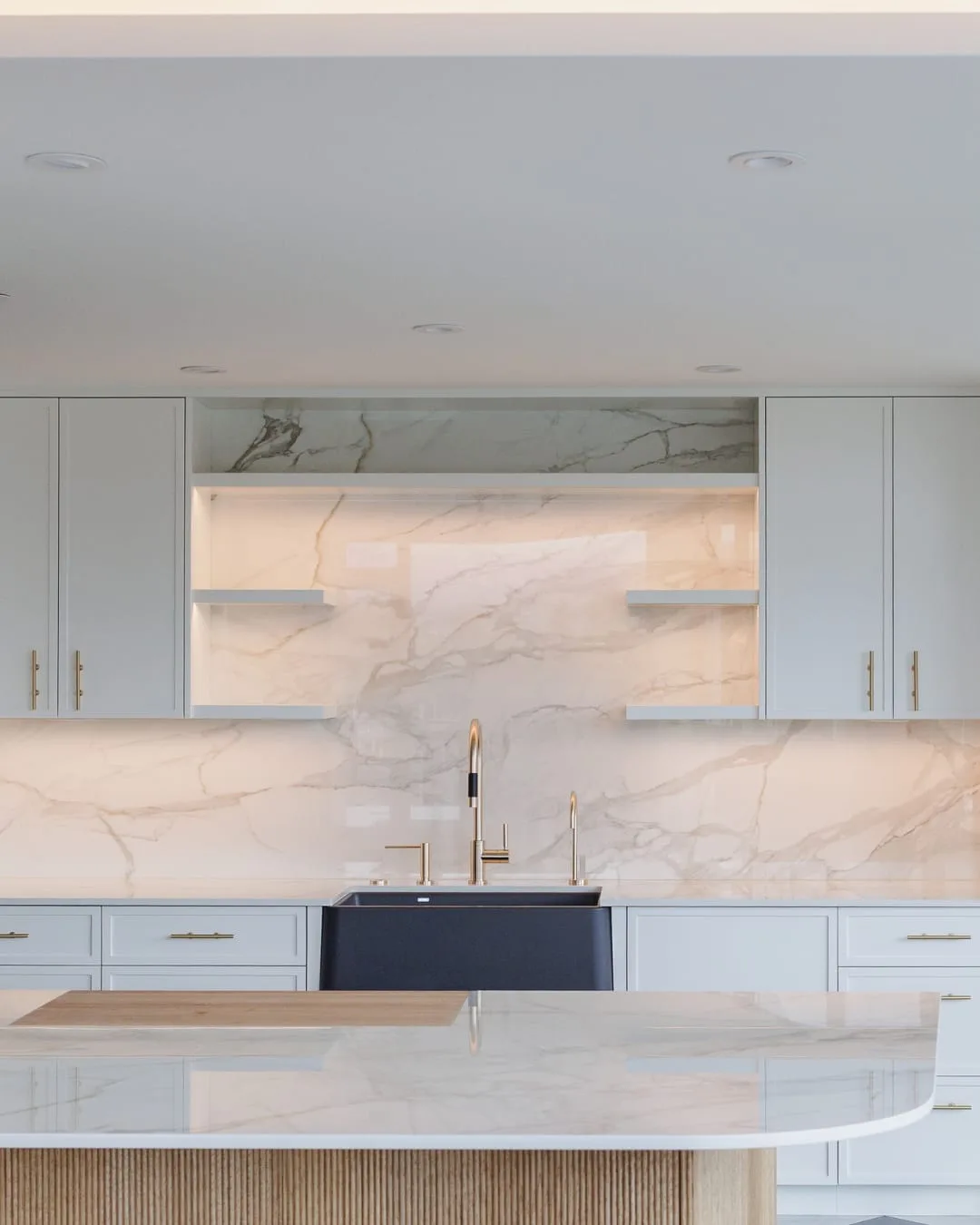 After: the rebuilt kitchen, with white cabinets, a marble slab backsplash with lit open shelves and a curved marble island.